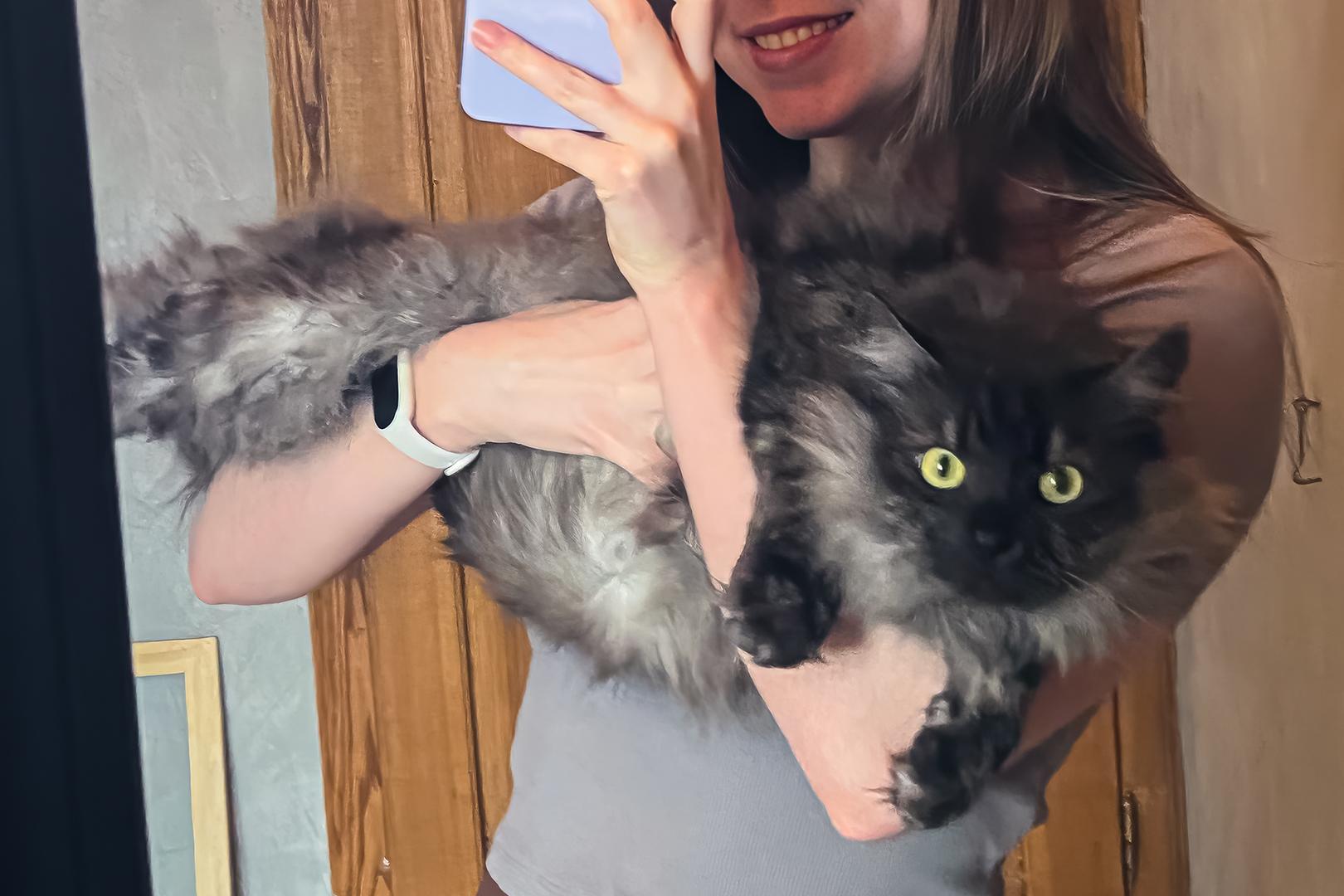 Photo of Baton, the dark grey long-haired cat, being held by my girlfriend Anna as she takes a selfie in the mirror. His back legs are suspended in the air over Anna's right forearm, his front legs gripping her left inner elbow for support as she holds her phone up to take a selfie, and his fluffy, smoke-colored belly hangs in the air in front of her chest. Baton's piercing green eyes are staring right at the camera through the mirror as Anna holds him in an awkward horizontal position. Through the black-framed rectangular mirror some gray walls, a wooden door, and a wooden art frame sitting in the corner of the room can be seen in the background behind them. Most of Anna's body is cropped out of the Baton-focused shot, with only the tight gray t-shirt on her upper body, the lower half of her face, and her beautiful smile in frame.